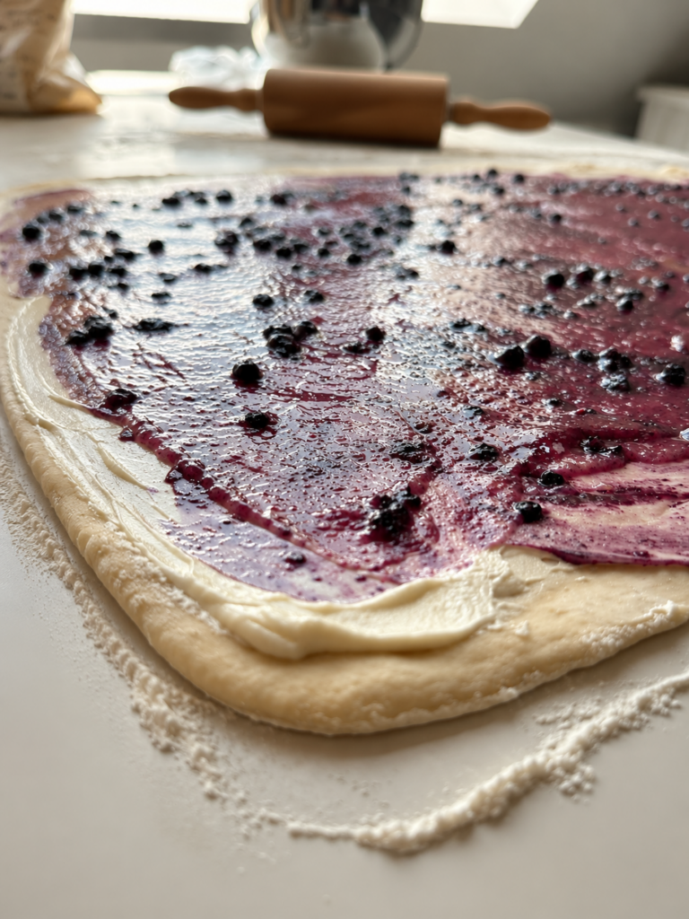 Nahaufnahme des Hefeteigs mit Frischkäse, Wildheidelbeerfüllung und frischen Wildheidelbeeren vor dem Aufrollen.